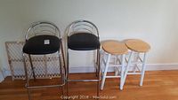 Four stools: two metal frame with black upholstered seats, two white wooden stools with round natural wood seats. They stand side by side on hardwood floor against wall.
