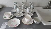 Over 10 delicate bone china cups, saucers, and plates displayed on countertop with various floral patterns visible.