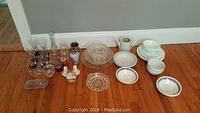 Various glassware including wine glasses, drinking glasses, vases, clear patterned bowls, white bowls and plates with rim accents, ceramic pitcher, decorative ceramic rooster figurine, and serving dishes.