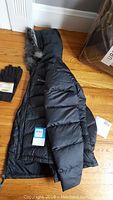 Black Columbia winter coat folded on the floor with fur trimmed hood visible and black gloves beside it.