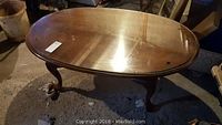 Wooden oval coffee table with glass top on curved legs, shown in a slightly worn basement setting.