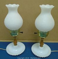 Pair of white milk glass hobnail table lamps showing full front view with scalloped glass shades, brass details, and wooden bases.