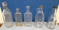Front view of five vintage glass bottles of various shapes and sizes on a light surface with a neutral background.