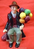 Front and left side view showing seated man holding colorful balloons, sitting on wooden crate, with red bag on ground.