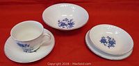 Photo showing one cup with saucer, two cereal bowls, and two bread and butter plates with royal blue floral design, scalloped edges, on red background