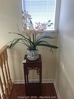 Full view of the plant stand with the potted artificial orchid on top placed near a window and stair railing.