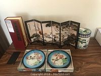 Full view of the lot showing the miniature two-sided screen, two framed circular mother of pearl art plates in boxes, and ceramic storage container with lid on wood surface.