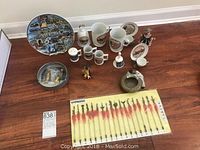 Photo showing the lot including miniature Asian weapons in display case, collectible plates from Vancouver and Victoria, ceramic mugs, small figurines, and ashtrays arranged on wooden floor.