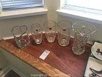 Five clear crystal vases displayed on a wooden ledge, including different shapes and sizes; heart-shaped vase on far left.