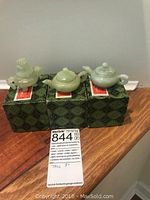 Photo showing three jade figurines placed on their original green patterned boxes with the auction label visible.