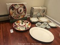 20-piece Menorca porcelain dinner set box with floral pattern plates, cups, and saucers, Ambrosia clay art serving platter with colorful vegetables and fruits, and large white serving platter on floor.