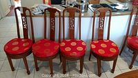 Front view of 4 wooden Asian-themed dining chairs with red cushions patterned with gold circles arranged side by side on tile floor in kitchen area.