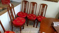 Four wooden dining chairs with carved Asian design backs and round red cushions with gold circular medallion patterns, some staining and leg damage visible.