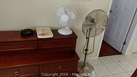 Side view of the dresser top showing a black radio, a rectangular clock radio, white table fan, and Noma floor fan standing on the floor near a wall.