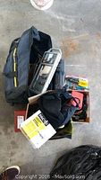 View of various tools in black soft-sided bag and cardboard box including tool belt, pliers, electrical tape, gloves, and other hand tools.