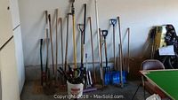 Wide view showing assortment of garden tools with long wooden handles, including shovels, rakes, and green-handled clippers arranged against a white wall along with a white bucket containing smaller tools and other implements.