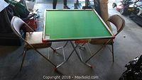 Card table with green playing surface and two metal and vinyl folding chairs in garage setting.
