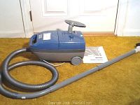 Full view of the blue Kenmore canister vacuum with attached hose and wand against a door and carpet background. Owner's manual visible on the floor.