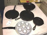 Set of five cast iron cooking pans arranged on a cloth surface: includes round skillet, two flat griddles, a muffin pan, and an oval omelet pan.