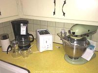 View of all appliances together on yellow countertop showing coffee pot, toaster, mixer, coffee grinder, and tea steeper.