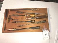 Nine antique soldering irons with wooden handles mounted on a pine wood display board.