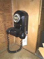 Full view of vintage black rotary dial wall phone mounted on wooden panel, handset hanging on hook with coiled cord.
