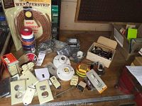 Overview of assorted electrical parts on table including outlet covers, light sockets, fuses, tape rolls, and metal junction boxes.