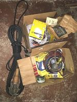 Photo showing black extension cord and trouble light with protective cage, multiple boxes containing GE Soft White 60 watt light bulbs and wires, electrical receptacles, and telephone parts.
