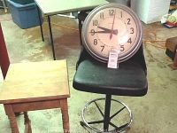 Vintage shop stool with black cushioned seat and Simplex wall clock on chair, antique side table in background