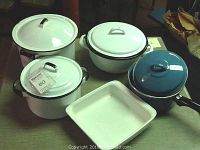 Five enamelware pots and a Corningware square baker arranged on the floor: a white stock pot with lid, white 5 qt. pot with lid, white 3 qt. pot with lid, blue enamel 3 qt. pot with lid, and a white Corningware square baking dish.