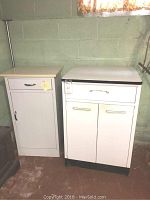 Two vintage metal base cabinets side by side. Larger cabinet with porcelain top on right, smaller cabinet with Formica top on left. White finish with metal handles.