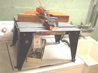 Full view of the metal Sears Craftsman router table with orange and black colors and accessories on top, placed on a white surface.
