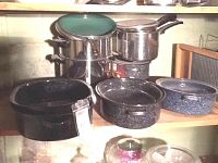 Photo showing a collection of stainless steel pots and blue speckled enamel roasters on wooden shelving.