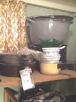 Large cast iron pots and a yellow enamel saucepan stacked on a wooden shelf with a green glass bowl and ceramic bowls.