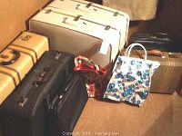 Collection of vintage luggage including white Featherlite suitcases, black vinyl cases, beige vinyl cases, and floral handbags on a carpeted floor under warm lighting.
