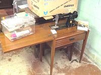 Photo showing the Singer sewing machine mounted on a wooden cabinet with a drawer and extended leaf, alongside a clear plastic sewing box containing sewing supplies and a wooden bench.