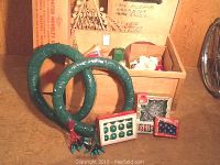 Photo showing two green wreath frames wrapped in tape, box of green glass bulbs, several smaller boxes of red and multi-colored small glass bulbs, and strands of blue vintage Christmas lights.