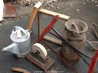 Wide view of lot showing galvanized watering can, milk can, two-person crosscut saw, wooden framed screened tray, grinding wheel, and other tools on asphalt.