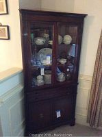 Front view of wooden corner hutch showing glass doors with dishes inside, drawers, and cabinet door below.