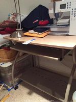 Photo of small desk showing wood laminate top, metal legs, keyboard drawer and foot shelf, items on desk not included except lamp.