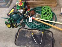 Photo showing assortment of garden tools and supplies laid out on a floor including hoses, watering can, sprayers, rakes, shovel, and a shoe tray.