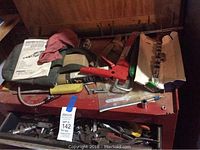 Top view of red tool chest showing various tools and hardware items including a caulking gun, clamps, screws, a screwdriver, and an instruction manual placed on top of the chest.