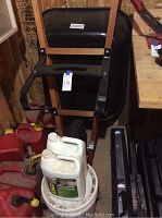 Photo showing black wheelbarrow leaned against a wall, a white bucket on the floor, and two white plastic containers placed on the bucket. Red fuel containers and other items are visible in the background.