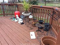 Corner view showing multiple garden decor items on a wooden deck including a red garden stool, plant stands, decorative statues, hanging stained glass lamp, and various plant pots and accessories.