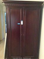 Front view of tall, dark wood armoire with closed double doors showing decorative molding and deep wood finish.