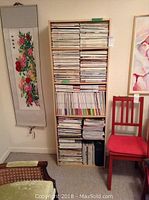 Photo showing the taller wooden bookcase filled with many magazines and papers, next to a red chair and a floral hanging scroll on the wall.