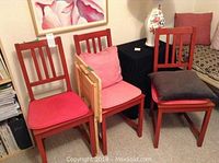 Three red painted wooden chairs with vertical slat backs and assorted cushions in red, pink, and gray colors. Chairs show minor paint nicks. Small natural wood TV table with folding sides and a natural finish next to the chairs.
