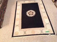 Full view of the smaller carpet with black center and beige border with symbols and a visible green label.