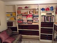 Wide view of two white laminated shelving units filled with books, binders, and decorative items, next to couch with cushions.