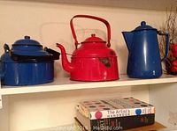 Three vintage metal enamel pots on a white shelf, showing red and blue colors, dents and wear.